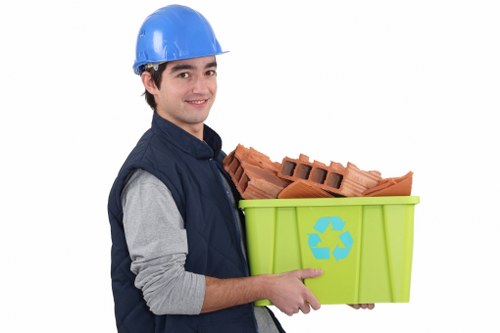A young male waste collection worker wearing a blue safety helmet and a navy work jacket over a grey long-sleeved shirt is holding a bright lime green recycling bin with a blue recycling symbol on its side. Inside the bin, there are several red clay bricks, stacked neatly and partially visible, with rough textures and rectangular shapes. The background is plain white, emphasizing the worker and the bin, with no environmental context shown. The worker is smiling and looking slightly to the viewer's right, suggesting a friendly and professional attitude. The image relates to rubbish removal services, highlighting the handling of construction debris or builder waste, typical for garden clearance or construction site waste collection in the Hatch End area. The overall composition is clean, professional, and focused on waste transportation, supporting local SEO for garden clearance and rubbish removal services.