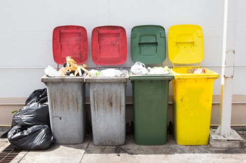 Four large wheelie bins with closed lids are lined up against a white wall, each designated for different types of rubbish. From left to right, the bins are black, grey, green, and yellow. The black and grey bins contain visible rubbish including plastic bags, paper, and miscellaneous waste, which are slightly overflowing. The green and yellow bins also hold waste, with some items protruding from the open lids, indicating recent collection or ongoing filling. To the left of the grey bin, there is a pile of tightly tied black rubbish bags on the pavement. The scene is set outdoors on a concrete surface, with a metallic grate visible adjacent to the black bags, and a narrow drainage channel beneath the bins. The setting suggests a typical residential area, likely near a property in Hatch End or surrounding part of northwest London. The image portrays a straightforward waste collection setup consistent with local rubbish disposal and garden clearance services offered by Garden Clearance Hatch End, emphasizing organized waste segregation and disposal options for households in the area.