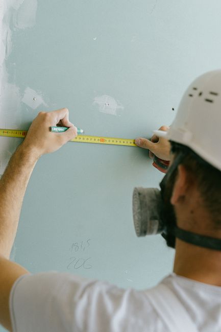 A construction worker wearing a white safety helmet and a protective mask is measuring a light blue wall with a yellow tape measure. The worker's left hand is holding the tape at one end, while the ri