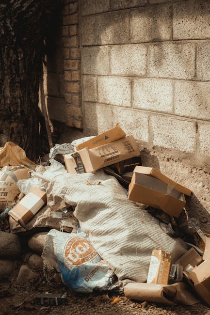 A collection of overflowing rubbish bins and scattered waste materials situated on a paved urban parking area. The pile includes a large grey mixed waste container filled with paper, cardboard boxes,